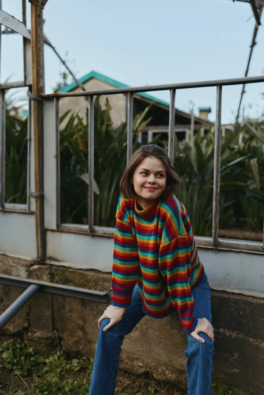 Woman in her mid 20s wearing a rainbow sweater and denim jeans, smiling and leaning on a greenhouse railing. Curly hair, casual outfit, cozy colorful fashion lifestyle portrait.