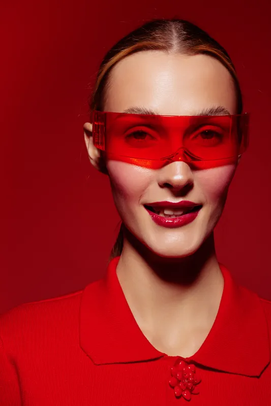 Portrait of a smiling woman wearing red transparent glasses and a red top against a red background. Concept of fashion, style, and modern accessories with vibrant color contrast.