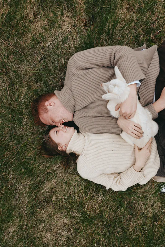 Couple outdoors on grass, cuddling a white goat kid, warm knits, intimate moment, gentle breeze, soft natural light, relaxed lifestyle, nature backdrop, shared affection