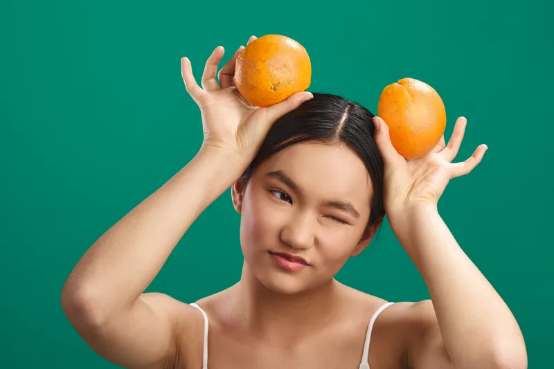 Asian woman playfully balances oranges on her head against a vibrant teal backdrop, conveying fun energy and creative styling for lifestyle photography.