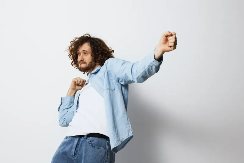 Young man with curly hair dancing joyfully in a casual light blue outfit against a plain white background, expressing happiness and carefree emotions