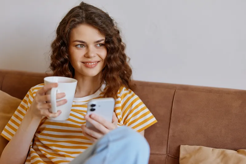 Young woman with curly hair sits on a sofa, sipping coffee and checking her phone in a cozy living room, expressing a calm, everyday moment at home.