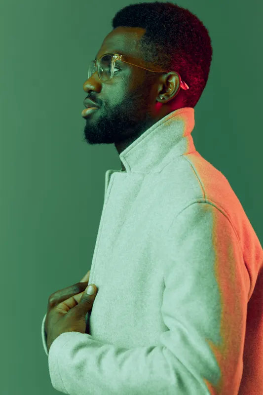 Portrait of a stylish Black man wearing a modern grey coat with a thoughtful expression against a colorful background featuring green and orange hues