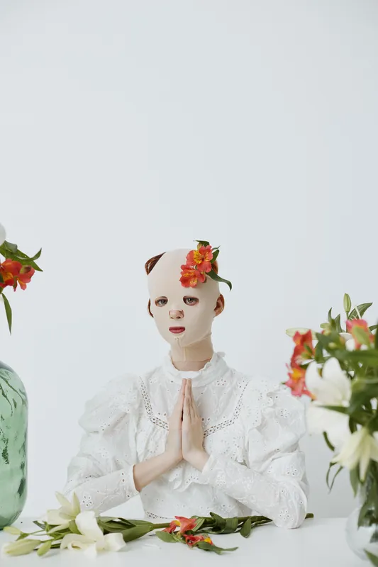 Artistic portrait of a masked model dressed in white, surrounded by flowers, conveying tranquility and serenity in a bright, minimalist setting