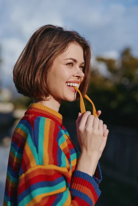 Woman mid 20s smiling in colorful striped sweater, holding yellow sunglasses by mouth, bob haircut, outdoors in sunlit urban park, playful lifestyle portrait for fashion and travel.
