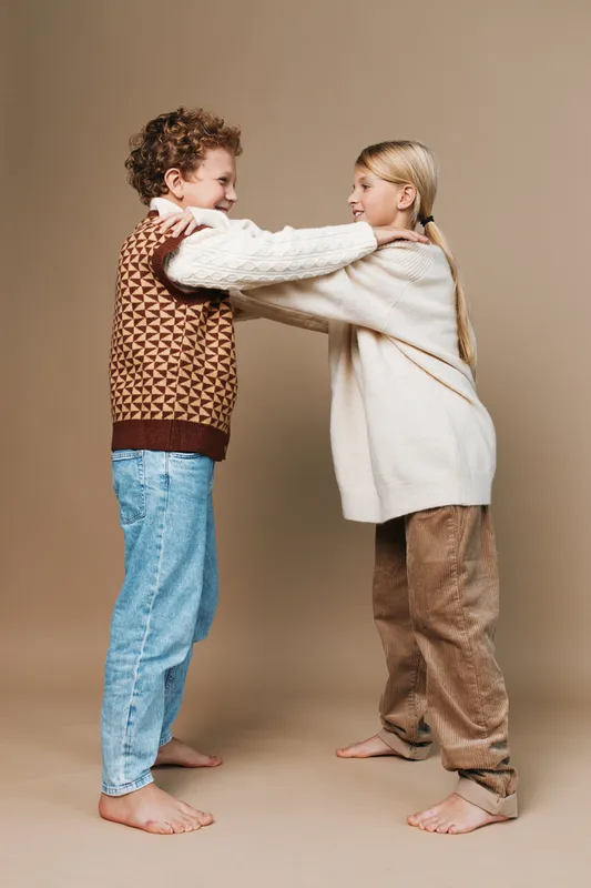 Two children share a warm hug in a soft studio setting, showing friendship, comfort, and playful connection through cozy knitwear and casual, relaxed pose.