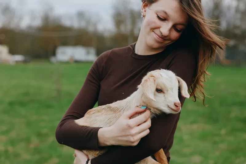 A woman in a dark sweater holds a young goat in a sunny field, smiling softly. A gentle moment of bond between human and animal, highlighting simple countryside life and care.