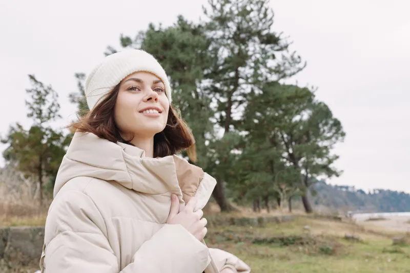 Woman in a beige winter coat and white beanie stands outdoors among trees, smiling softly, exuding cozy style and serene confidence in natural light