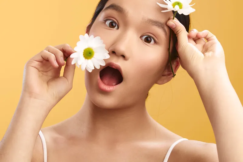 asian woman with a surprised expression holds flowers against a warm yellow background, creating a fresh, playful portrait that radiates brightness and charm.