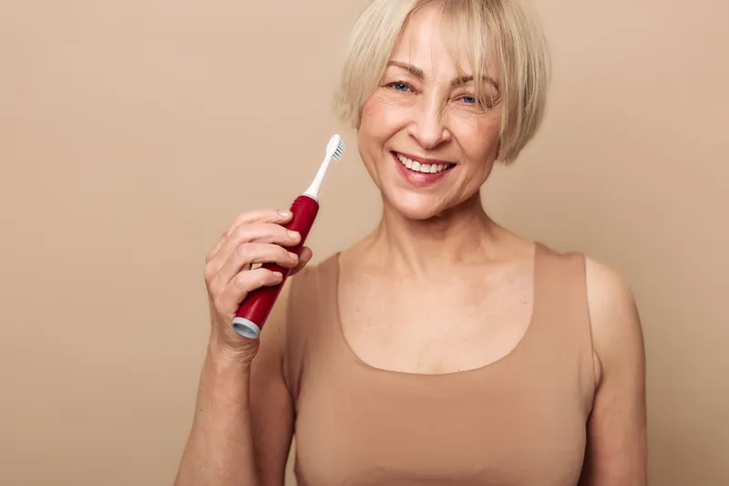 Smiling mature woman with short blonde hair holds electric toothbrush near face on beige background. Concept of dental hygiene, oral care, personal routine, and healthy lifestyle for seniors.