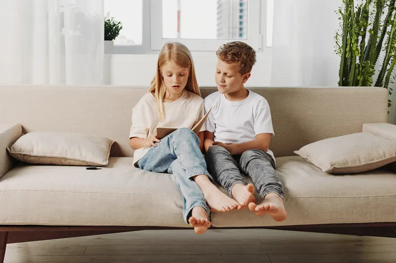 Two children sitting barefoot on beige couch reading a book together in bright living room with natural plants near window.