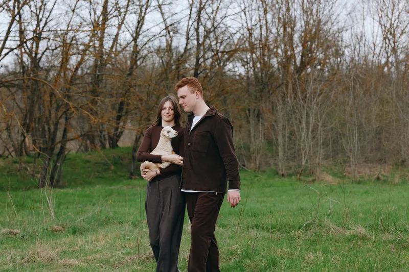 Lifestyle couple walks outdoors in park, woman holding small dog, man beside her, calm affectionate moment, natural light, green grass, bare trees in background, cozy casual mood.