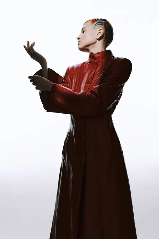 Woman with colorful braided hair wearing a long red leather coat posing gracefully against a bright white background, showcasing fashion and artistic style.