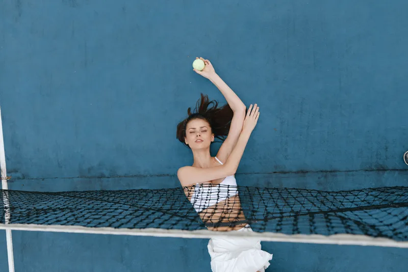 Young woman playing tennis with a ball against a blue background, showcasing athleticism and focus while wearing a sporty outfit