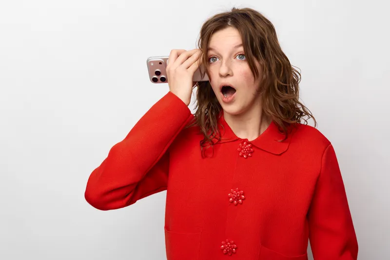 Surprised young woman in red sweater holds smartphone to ear, expressing shock or disbelief. Isolated on white background, concept for communication, reaction, and unexpected news.