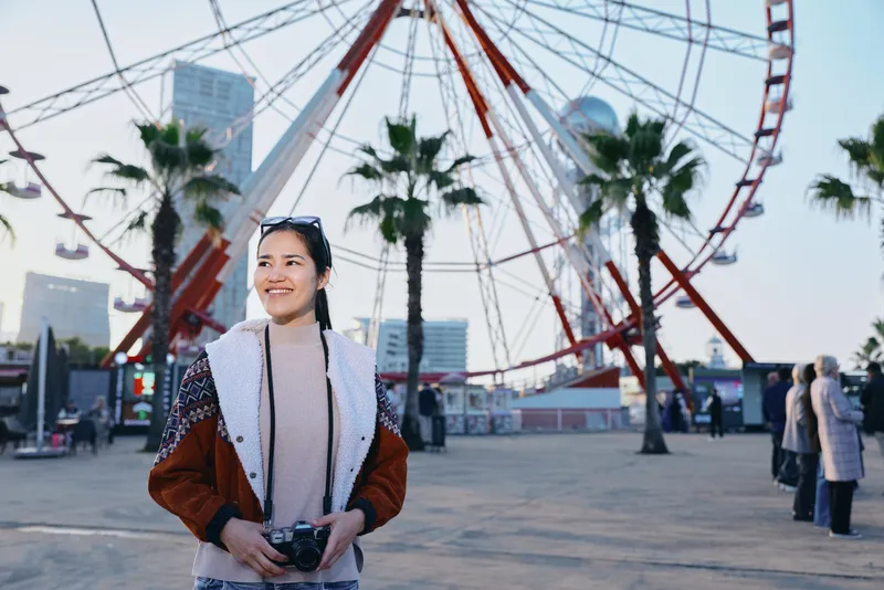 Smiling woman with a camera in a sunny outdoor amusement park, ferris wheel and palm trees in the backdrop, casual jacket, candid lifestyle moment, urban plaza setting, travel vibe, cheerful scene