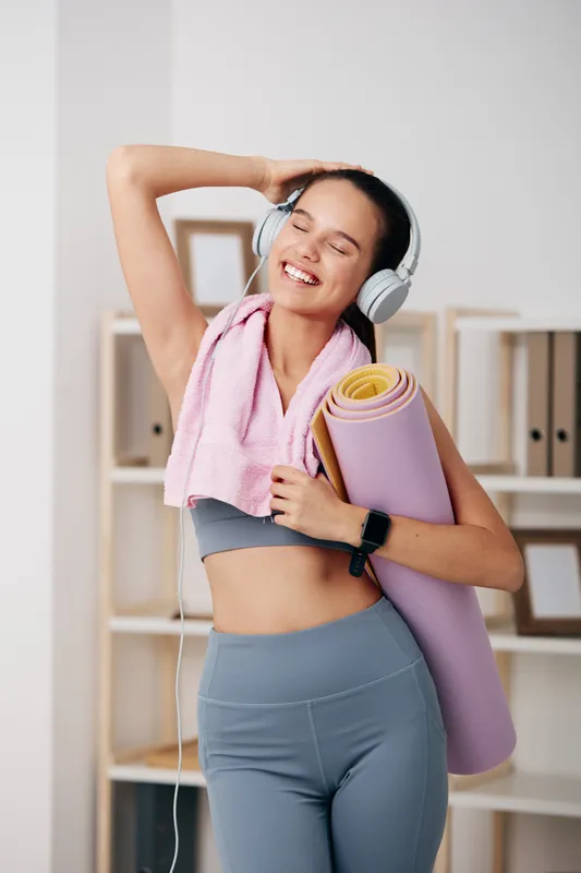 Female fitness enthusiast in active wear, wearing headphones and carrying a yoga mat, smiling while stretching in a bright home gym setup.