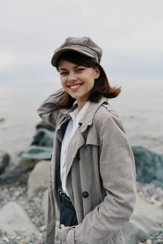 Smiling young woman wearing a patterned coat and cap stands by rocky shore, looking relaxed and happy. Outdoor portrait with natural light and casual autumn fashion style.