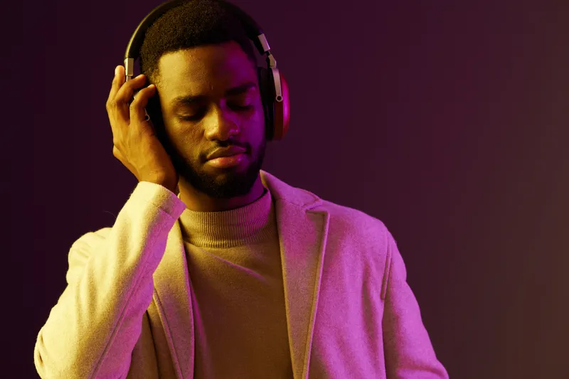 A young Black man wearing headphones, enjoying music, dressed in a stylish sweater and coat, set against a vibrant purple background showcasing a trendy urban vibe