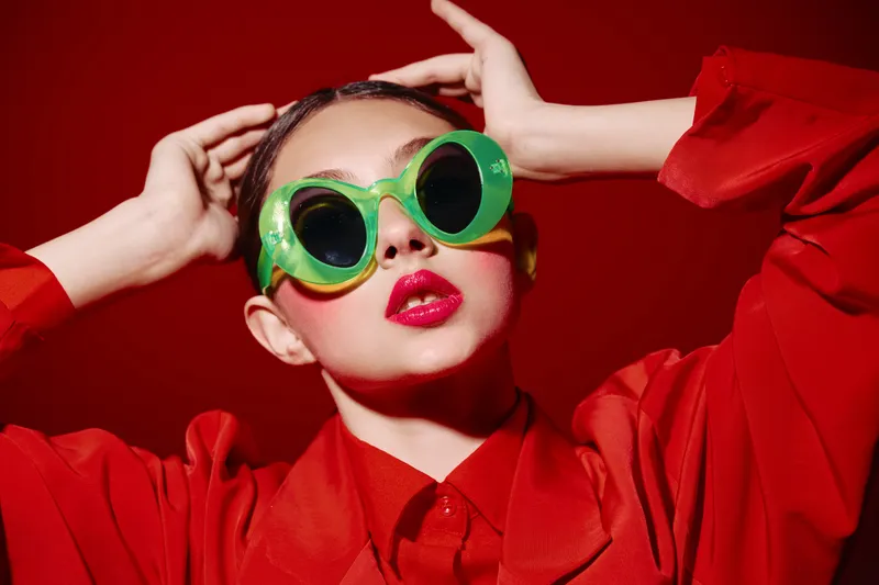 Stylish woman in a red blouse wears oversized green sunglasses and bright red lipstick. Studio shot with vivid red background. Concept for beauty creative fashion avantgarde style.
