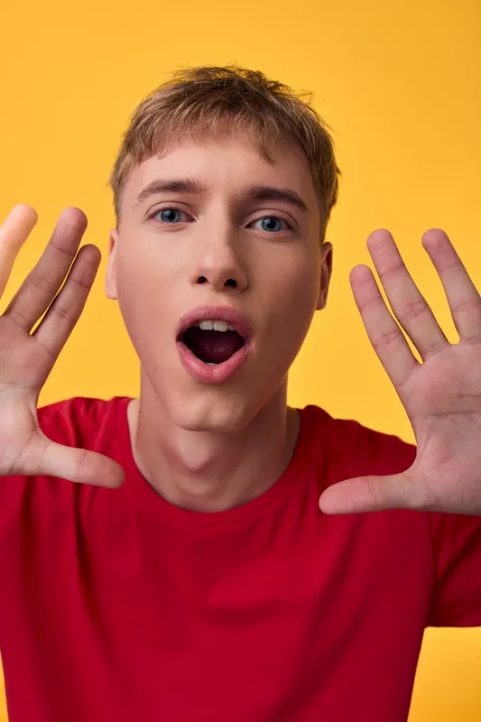 Surprised expression on a man with hands up against a yellow background, wearing a red shirt, capturing energy, excitement, and astonishment in a vivid closeup.