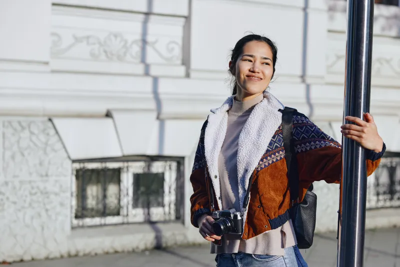 Woman with camera on a street outdoors, wearing layered cardigan and scarf, urban fashion vibe, candid street photography mood, natural light and confident expression in daylight