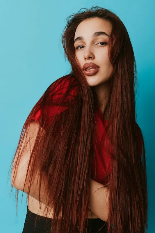 Woman with long straight brown hair and natural makeup posing with crossed arms against blue background. Fashion portrait with casual red top and confident expression in studio lighting.