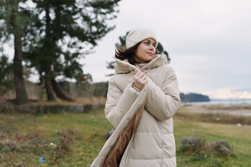 Woman in a beige puffer coat stands outdoors in a windy park, wearing a knit scarf and beanie, looking softly into the distance with calm expression.