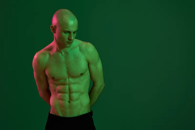 Muscular man with a pensive expression poses against a vibrant green background, showcasing his physique and confidence in a striking color light setting.