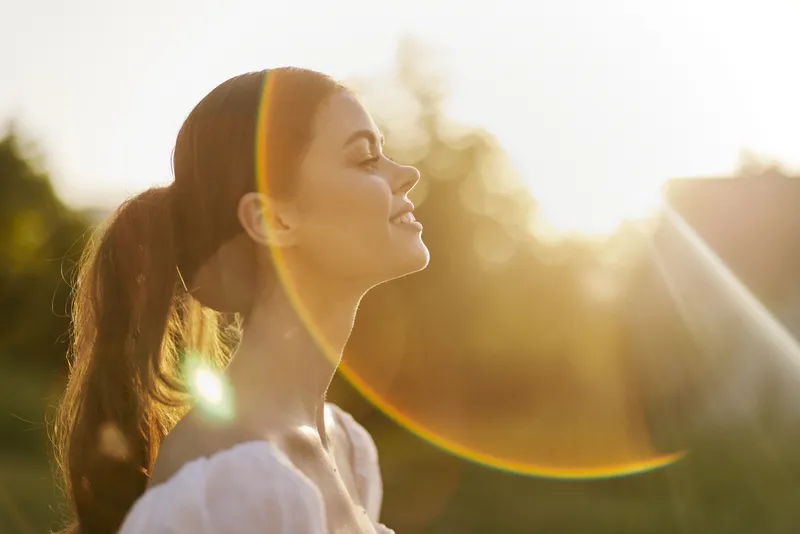 Young woman enjoying a sunny day outdoors, exuding happiness and tranquility in soft sunlight, with a lush green background and warm colors enhancing the serene atmosphere