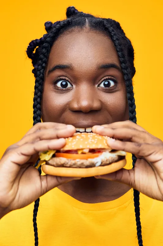 Happy woman with braids excitedly holding a burger, showcasing vibrant colors against a yellow background, emphasizing healthy eating and food enjoyment