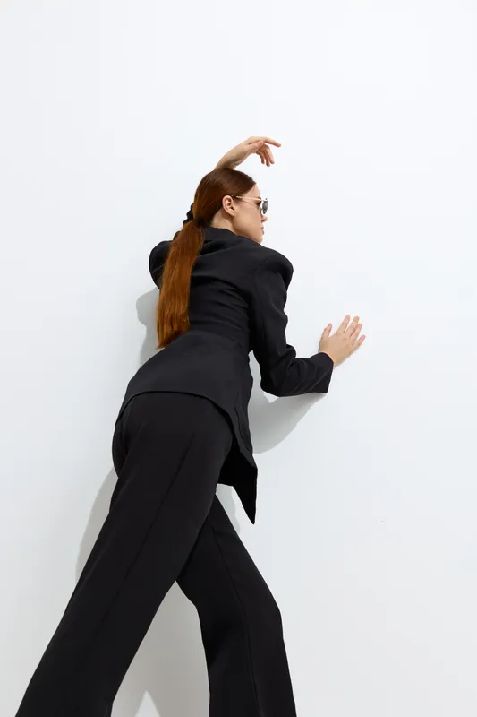 fashion woman in black suit poses stylishly against white wall, showcasing modern clothing and sleek hairstyle with sunglasses in minimalistic studio setting.