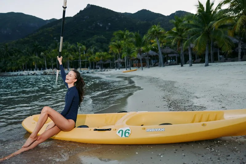 Young woman sits in a yellow kayak on a sandy beach, lifting a paddle as palm trees and distant mountains frame a serene coastal scene, calm water and soft light.
