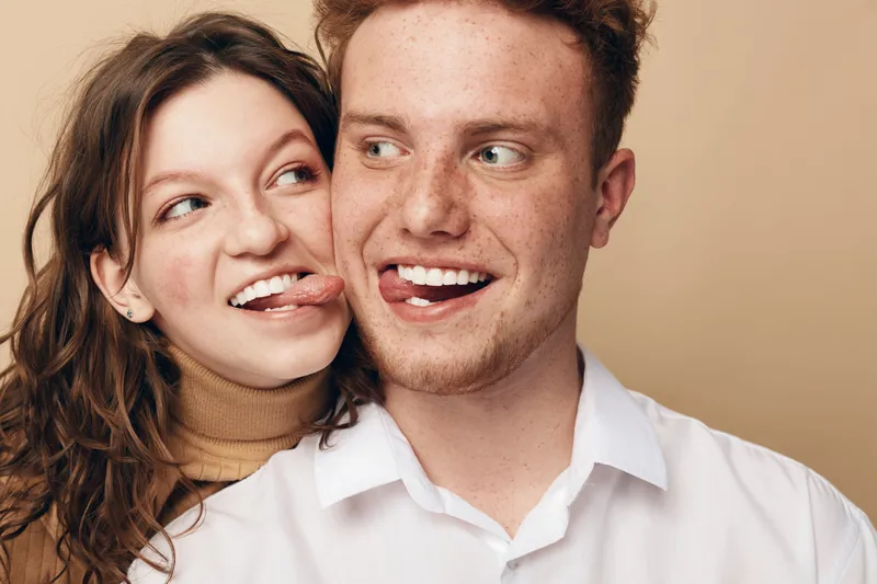 Young couple with playful expressions sticks out tongues, smiling and looking at each other. Casual clothing and warm beige background create a fun and intimate mood.