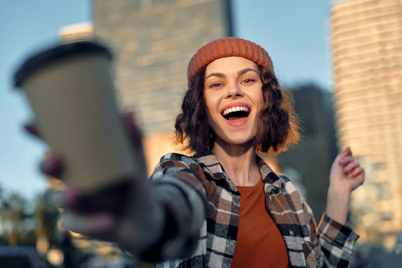 Woman holding coffee cup in city, beanie and plaid jacket, radiant smile and golden hour glow creating authenticity and candid lifestyle moment of mindful living and emotional storytelling.
