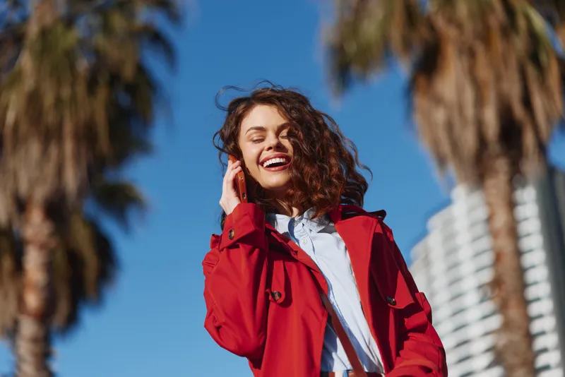 Young woman with curly hair laughs while wearing a bold red jacket outdoors, standing under a clear blue sky with palm trees in the background, conveying energy, confidence, and sunshine.