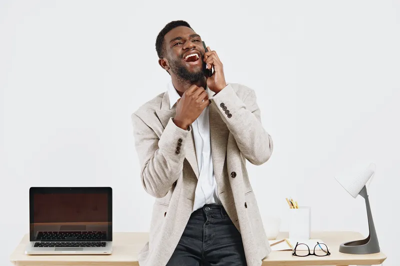 Full body joyful young African American man wearing stylish beige blazer and white shirt talking on smartphone isolated on plain light gray background studio portrait. People lifestyle concept