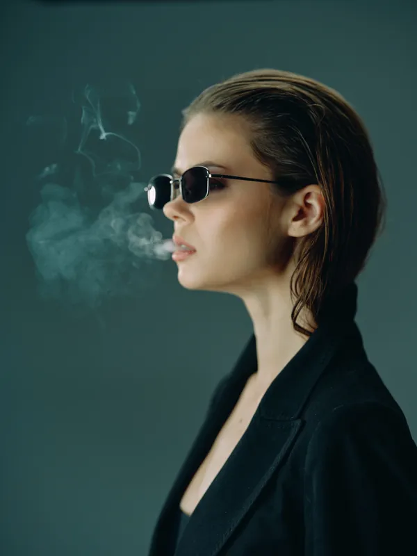 Woman with sunglasses smoking while wearing a black jacket in a dark setting with smoke around her.