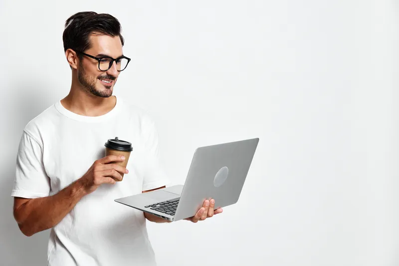 Smiling man in casual white t-shirt holding coffee cup while using laptop, looking at the screen with a positive expression, isolated on plain background.
