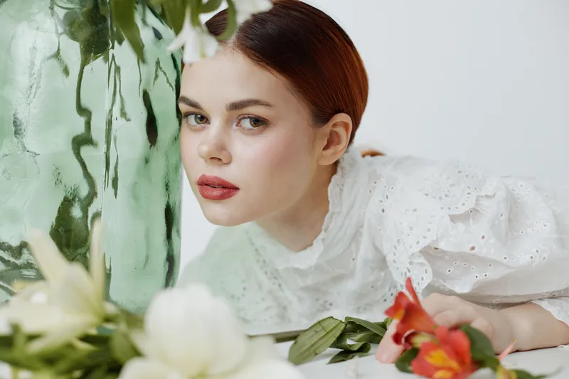 Young woman with elegant makeup resting her head on a green vase, surrounded by spring flowers, expressing tranquility and beauty in a bright, soft color palette