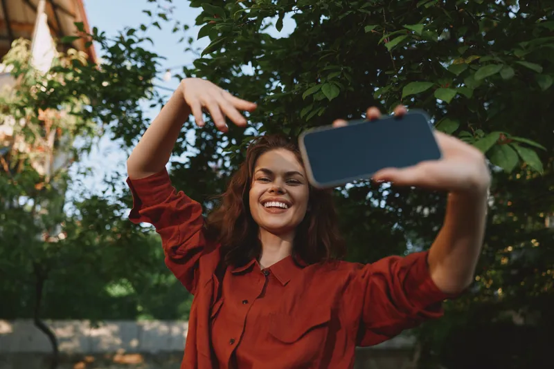 A smiling woman in a red shirt takes a casual selfie outdoors, surrounded by lush greenery in daylight. Portrait style captures confident, carefree moments