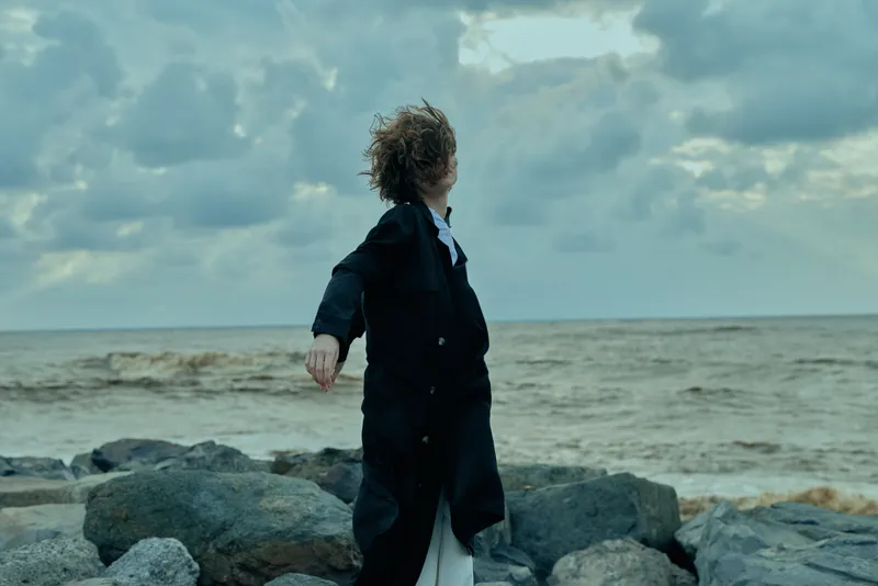 Wind swept figure stands on a rocky shoreline in a dark coat, gazing toward the sea under a dramatic cloudy sky, capturing motion and mood.