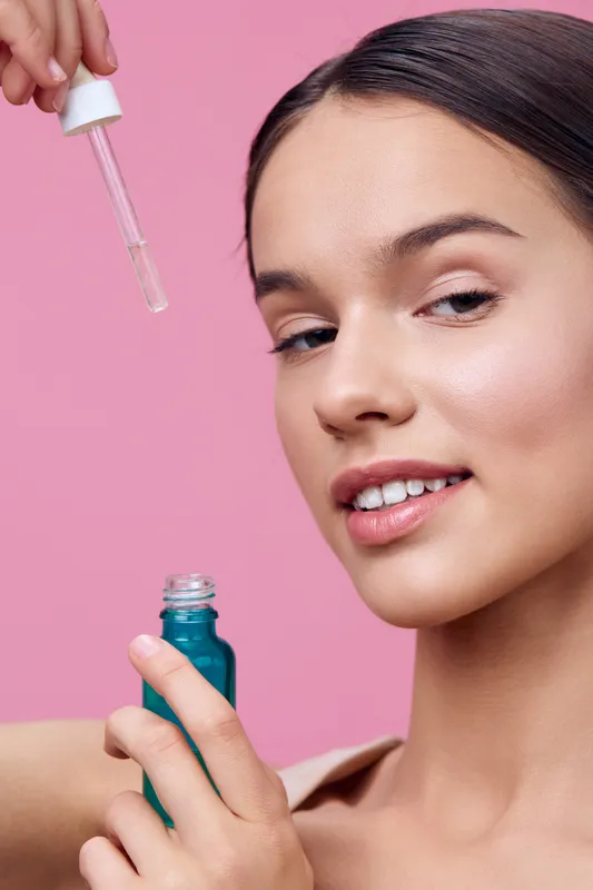 Young model applying skincare serum with a dropper against a pink background, showcasing healthy skin, beauty, and self care
