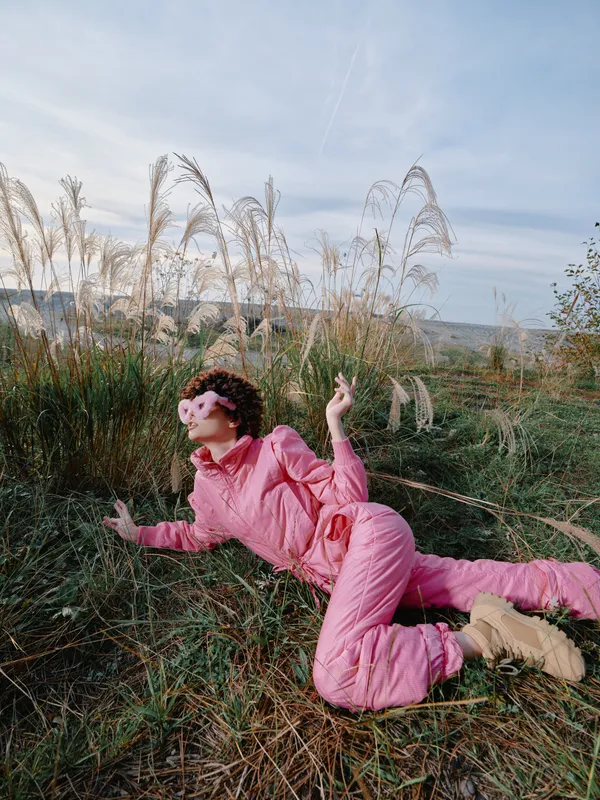 Person wearing pink jumpsuit relaxing outdoors on grass with tall plants, showcasing carefree style in a natural landscape and soft sunlight