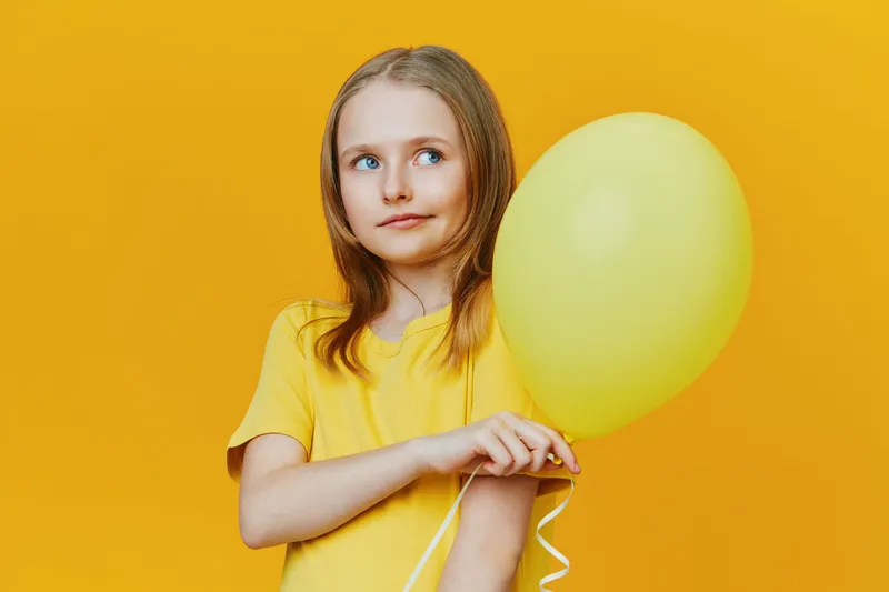 Young girl in a yellow shirt holding a yellow balloon against a bright yellow background, expressing curiosity and joy, perfect for seasonal concepts