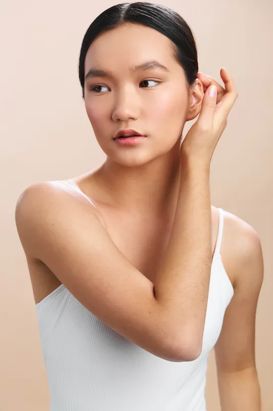 Asian woman regards the distance with a calm, elegant pose in a simple studio portrait, expressing serenity, focus, and understated confidence in soft light.