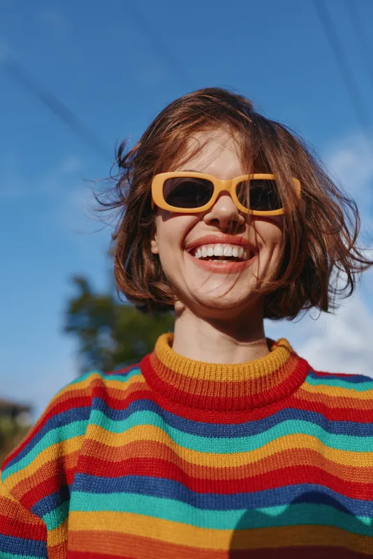 Woman with smile, sunglasses and rainbow sweater in a portrait shot, happy short hair model outdoor in sunny fashion scene, colorful casual knitwear and joyful lifestyle vibe.