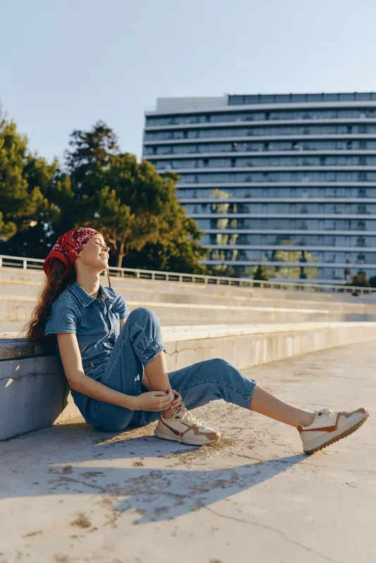 Lifestyle woman in modern boho-western denim street style sitting outdoors, enjoying sunlight with casual sandals and a red bandana, film color vibe.
