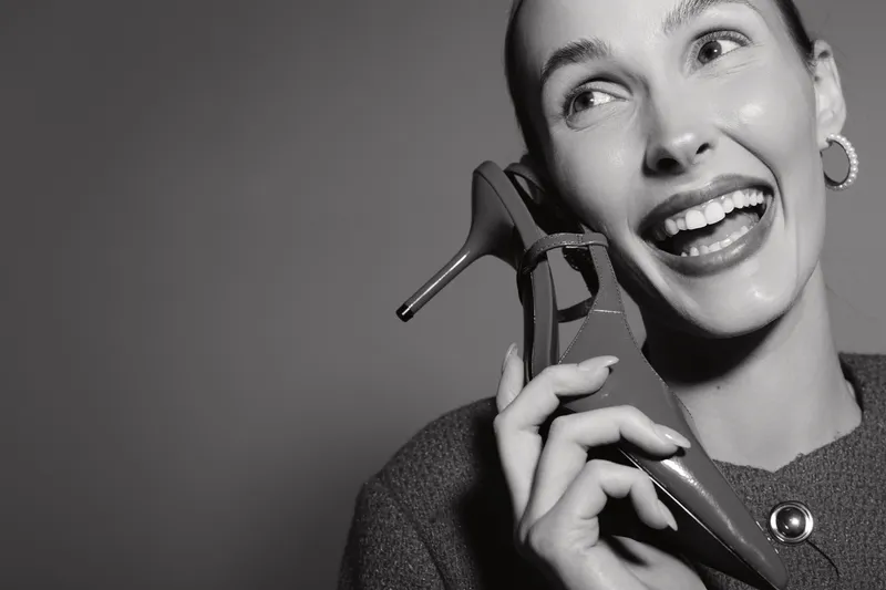 Happy Caucasian woman with light skin holds high heel shoe near face smiling in studio. Concept of fashion, joy, elegance, and lifestyle in black and white photography.