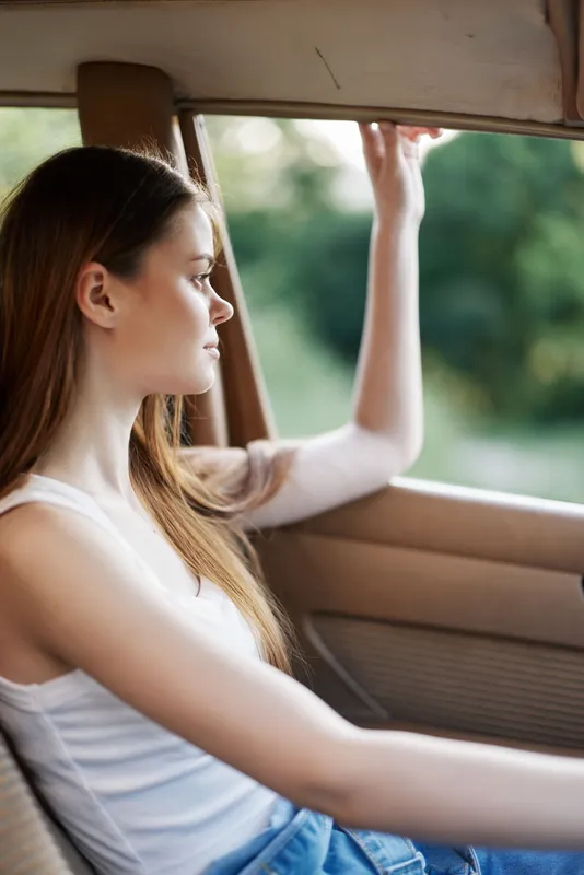 Young woman enjoying a beautiful day, gazing out of a vintage car window, wearing a casual white tank top and denim jeans, surrounded by greenery.